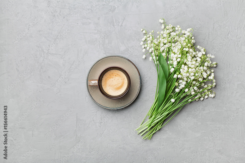 Coffee mug with bouquet of flowers lily of the valley on gray stone table from above in flat lay and minimalistic style. Beautiful morning breakfast. Coffee mug with bouquet of flowers lily of the valley on gray stone table from above in flat lay and minimalistic style. Beautiful morning breakfast.