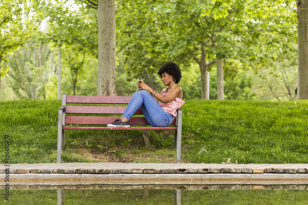 Fototapeta premium portrait of a Happy young beautiful afro american woman sitting on a bench and listening to music on her mobile phone. Green background. Lifestyles. Casual clothing.