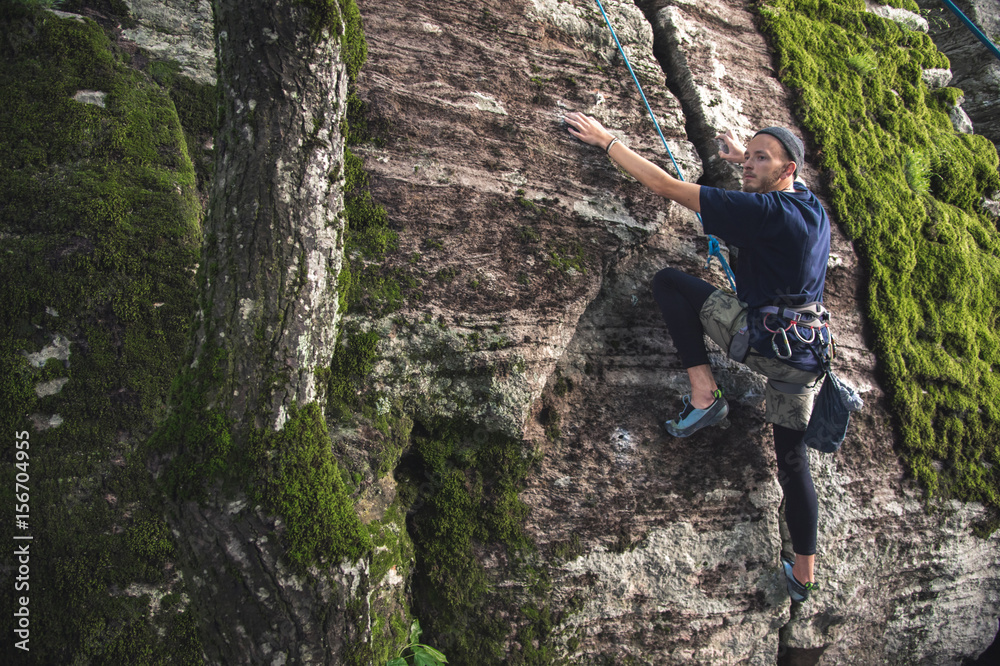 A young hipster is engaged in rock climbing with insurance on rocks with green moss