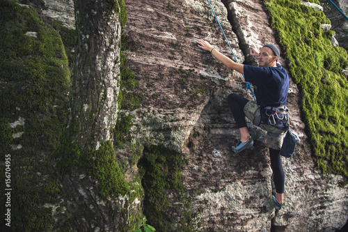 A young hipster is engaged in rock climbing with insurance on rocks with green moss