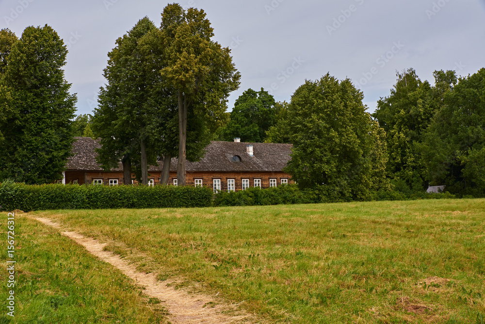 A merchant's house among the trees/Wooden merchant house surrounded by trees. The trees are high. In the foreground meadow and footpath. Russia, Pskov region, summer.