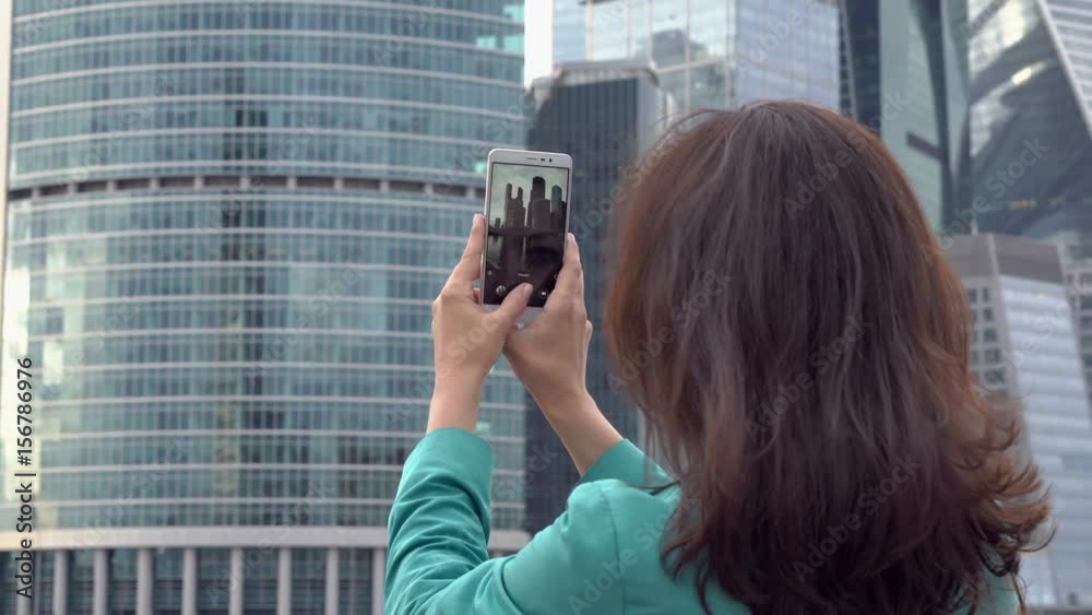 Cute tourist takes photos of the futuristic business center by camera of the smartphone. On the screen of the mobile device you can see the reflection of her eyes and face, close up UHD 4K Cute tourist takes photos of the futuristic business center by camera of the smartphone. On the screen of the mobile device you can see the reflection of her eyes and face, close up UHD 4K