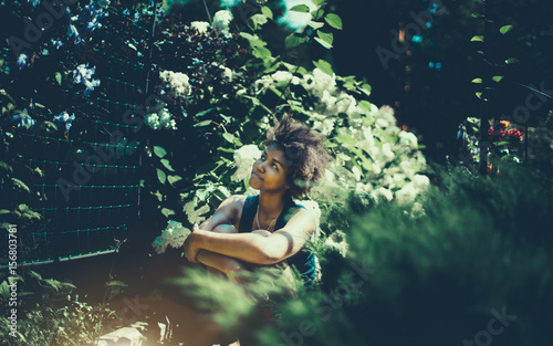 Fototapeta Naklejka Na Ścianę i Meble -  Attractive black curly girl is sitting in beautiful spring garden and looking on light blue flowers on fence, cute teen afro american female enjoying her leisure in private summer park on sunny day