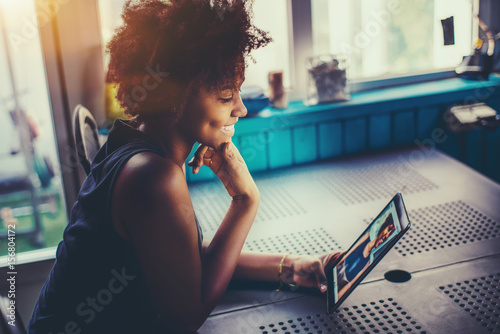 Laughing cute young black girl with perfect smile and dimples sitting at the kitchen table of summer house and making selfies to online chat with her friend using frontal camera of digital tablet photo