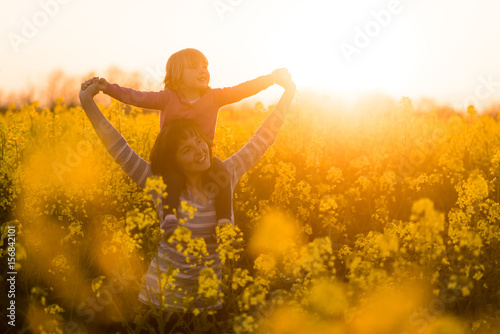 Mother carrying her little girl on shoulders, sunset background.