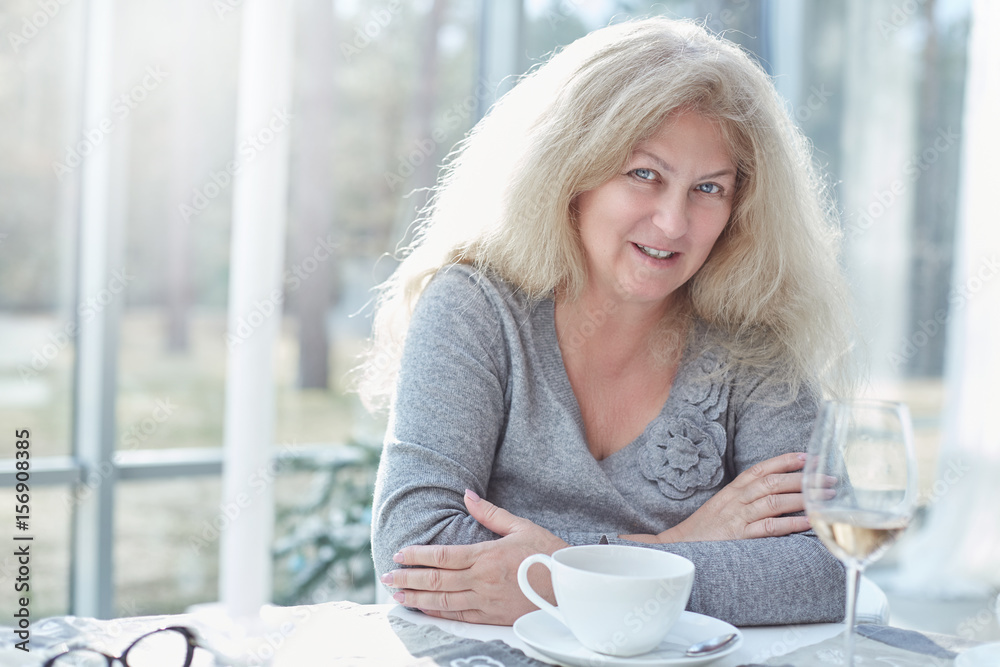 Close up portrait of beautiful blond mature woman looking at the camera cheerfully, having lunch in a beautiful light interior of luxury and stylish country house.Daily life activities concept. Close up portrait of beautiful blond mature woman looking at the camera cheerfully, having lunch in a beautiful light interior of luxury and stylish country house.Daily life activities concept.