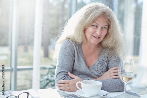 Close up portrait of beautiful blond mature woman looking at the camera cheerfully, having lunch in a beautiful light interior of luxury and stylish country house.Daily life activities concept.