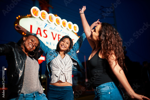 group of excited girls on vacation jumping for photo in front of welcome to las vegas sign at night