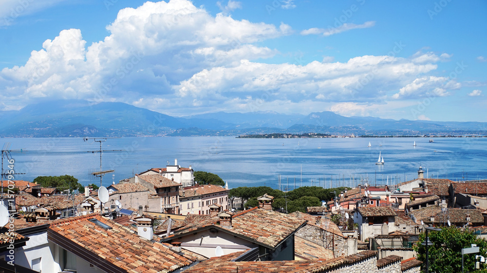 Amazing panorama from Desenzano castle on Lake Garda with old city roofs, mountains, white clouds and sailboats on the lake, Desenzano del Garda, Italy
