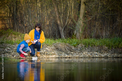 Portrait of woman with cute kid boy watching handmade toy boat sailing in the river. Mother with son having fun in the park on a spring or autumn day. Together. Lifestyle.