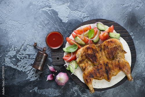 Roasted tapaka chicken served on tortilla bread with fresh vegetables. Flat-lay over grey stone background, horizontal shot photo