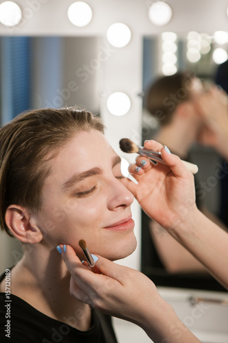 Men's makeup. A happy man will enjoy the make-up process. Make-up artist puts eye shadow and powder on face with brushes. Vertical indoors shot.
