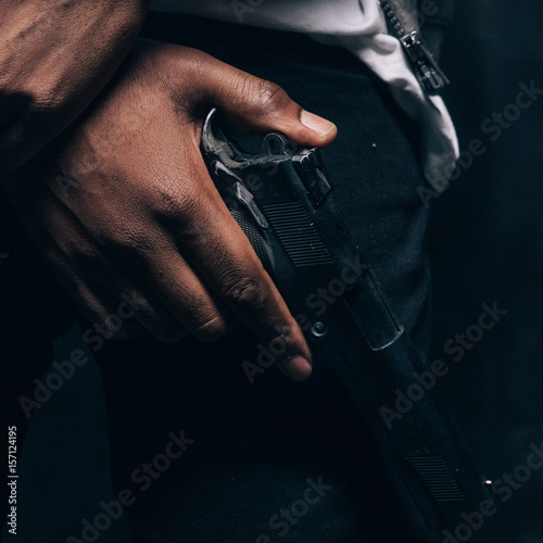 Unrecognizable armed black criminal man closeup studio shoot. Gangster guy with gun in hand on dark background. Outlaw, ghetto, murderer, robbery concept