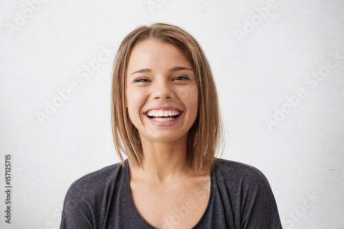 Positive human facial expressions and emotions. Cheerful attractive teenage girl with bob hairstyle grinning broadly, showing her perfect white teeth at camera while spending nice time indoors