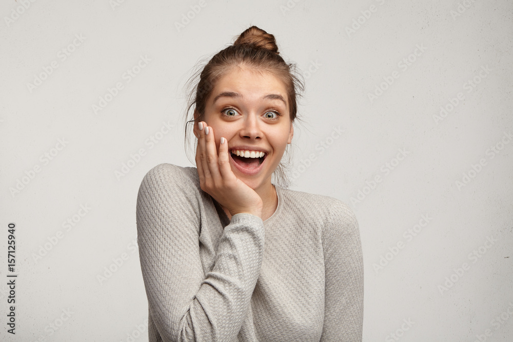 Beautiful charming young Caucasian 20 y.o. woman with hair knot opening mouth widely, saying Omg, Wow, having excited astonished look, holding hand on her face, delighted with unexpected gift Beautiful charming young Caucasian 20 y.o. woman with hair knot opening mouth widely, saying Omg, Wow, having excited astonished look, holding hand on her face, delighted with unexpected gift