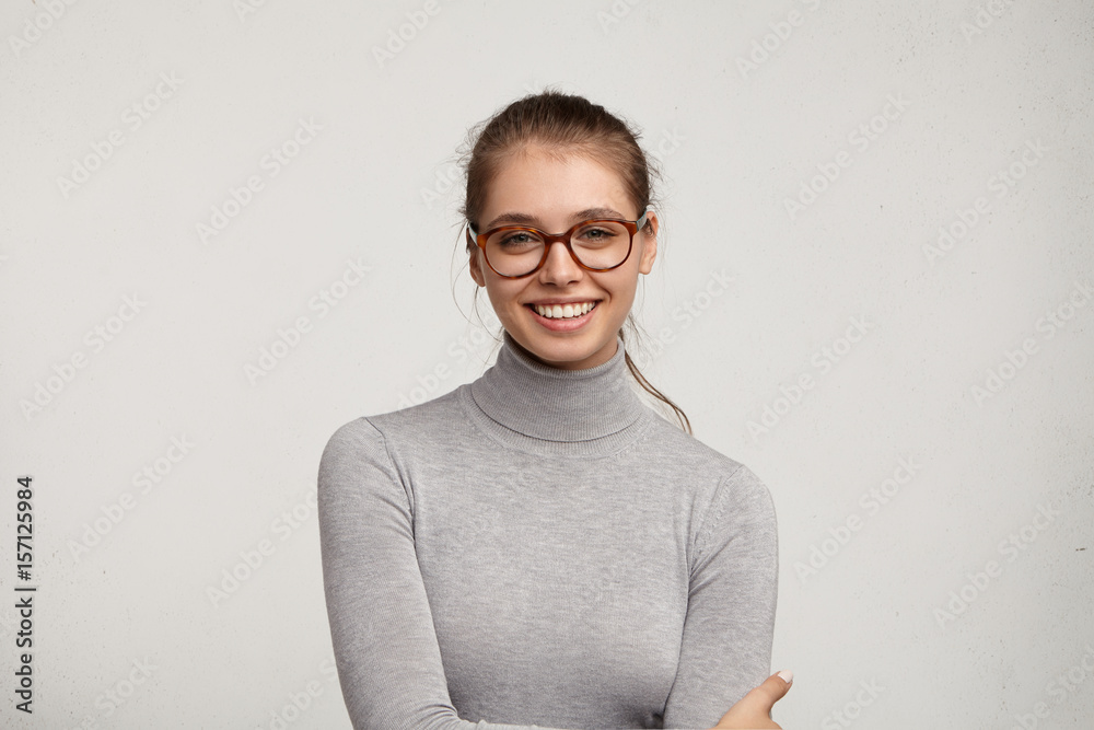 Foto de Headshot of cute student girl wearing grey turtleneck sweater and  stylish eyeglasses smiling broadly, having carefree and relaxed look while  resting at home after morning lectures at college do Stock