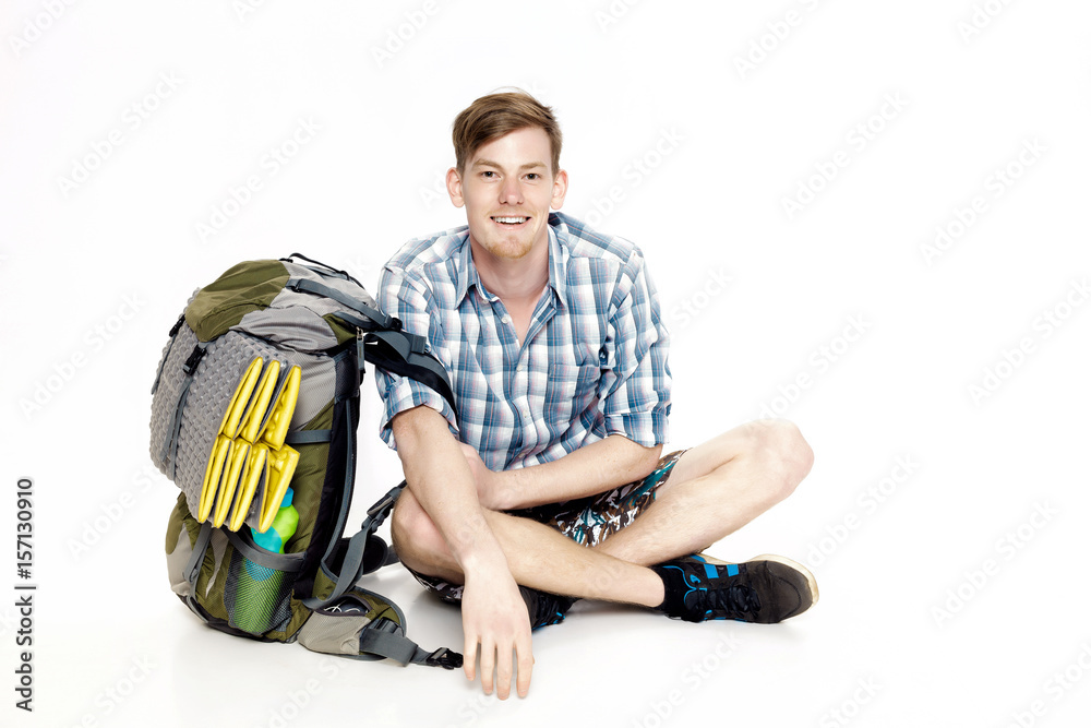 Young smiling tourist is sitting with backpack on white background. traveler is preparing for hike Young smiling tourist is sitting with backpack on white background. traveler is preparing for hike