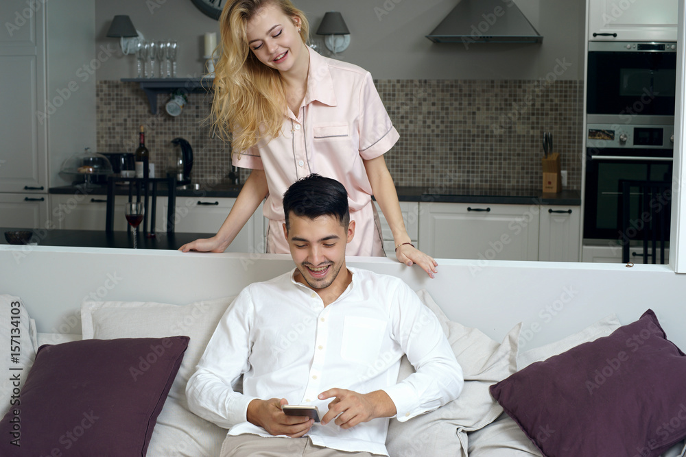 Handsome brunette man sitting at sofa and using smartphone. Pretty beautiful caucasian girl with bllonde hair standing from the back and looking what he do Handsome brunette man sitting at sofa and using smartphone. Pretty beautiful caucasian girl with bllonde hair standing from the back and looking what he do