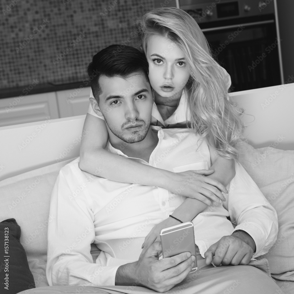 Handsome brunette man sitting at sofa and using smartphone. Pretty beautiful caucasian girl with bllonde hair standing from the back and looking what he do Handsome brunette man sitting at sofa and using smartphone. Pretty beautiful caucasian girl with bllonde hair standing from the back and looking what he do