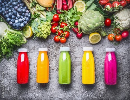 Healthy red, orange, green, yellow and pink Smoothies  and juices in Bottles on grey concrete background with fresh organic vegetables , fruits and berries ingredients, top view