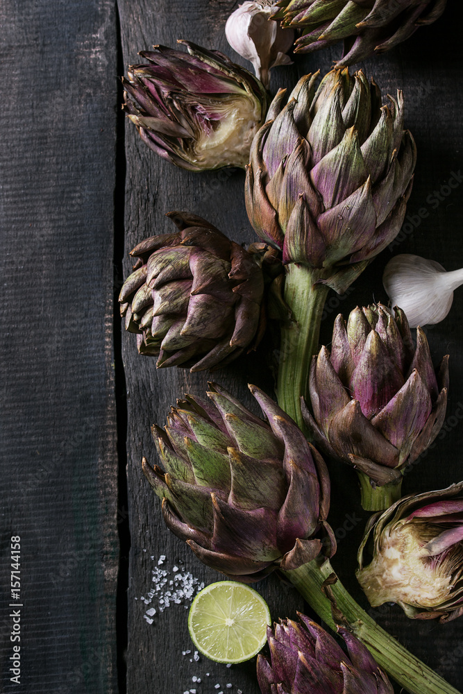 Obraz premium Uncooked whole and sliced organic wet purple artichokes with salt, lime and garlic over dark wooden background. Rustic style. Top view with space