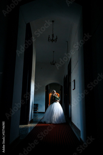 The bride in a dark large corridor stands at the window in her wedding dress, the girl in a bouquet on the red carpet