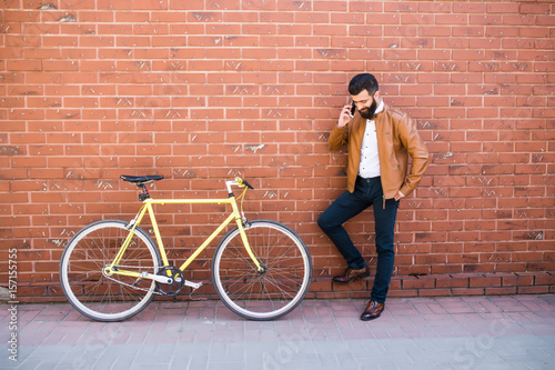 Young hipster man speak phone with his fixed bicycle on the wall