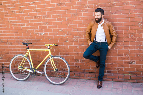 Young stylish man with beard in a brick background standing near the bike with bag in hands