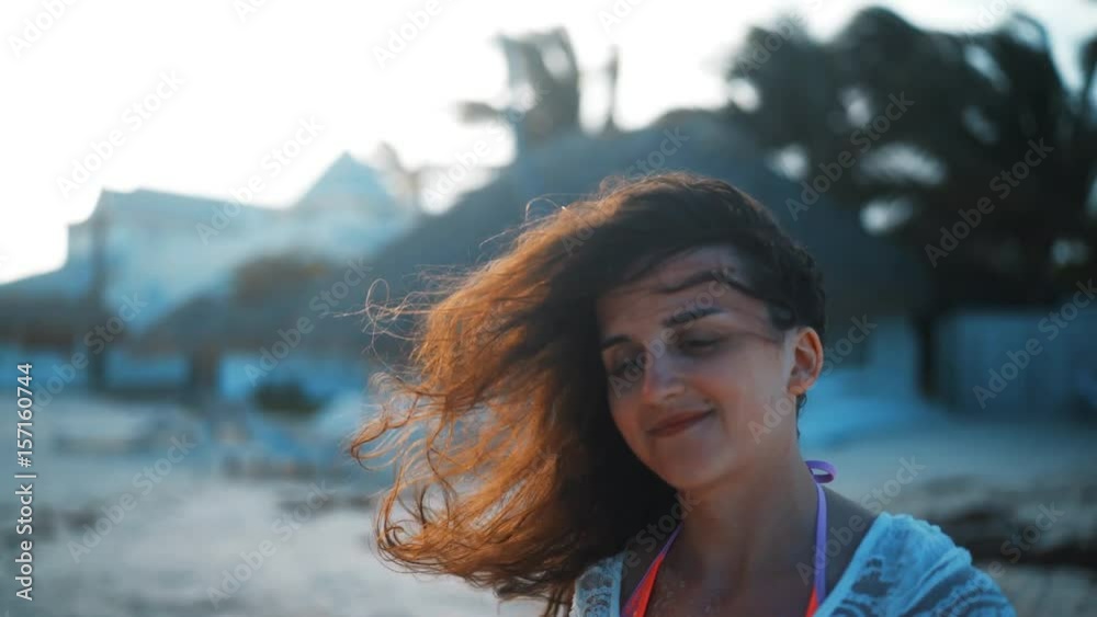 Close up portrait of young seductive brunette woman smiling on camera and laugh, hair blowing in wind. Sunny beach background at sunset. 4k ultra hd handy shot.