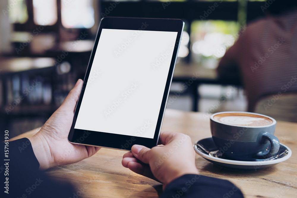 Mock up image of business woman's hands holding black tablet with white blank screen and coffee cup on wooden table in cafe Mock up image of business woman's hands holding black tablet with white blank screen and coffee cup on wooden table in cafe
