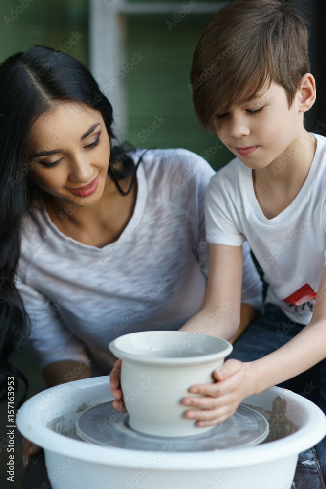Pottery classes. Young beautiful pretty caucasian brunette mother teaching her son. Son making clay por on potter wheel Pottery classes. Young beautiful pretty caucasian brunette mother teaching her son. Son making clay por on potter wheel