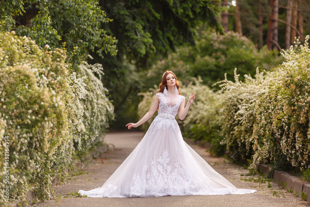Beautiful redhead Bride in fantastic wedding dress in blooming garden. Growth Portrait in sunset light. Pretty young caucasian redhead girl walks in a garden and plays with her dress. Young princess