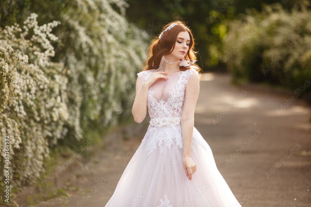 Beautiful redhead Bride in fantastic wedding dress in blooming garden. Portrait in sunset light. Pretty young caucasian redhead girl walks in a garden and plays with her dress. Young princess