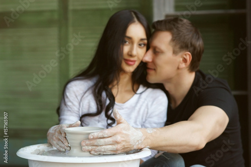 Romantic couple making clay pot on potter wheel. Handsome man hugging beautiful brunette woman. Shalllow DOF. Focus on man hand on pot.