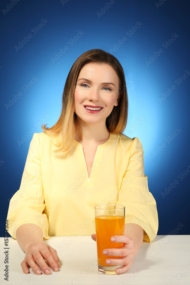 Beautiful woman sitting at table with glass of fresh juice, on color background Beautiful woman sitting at table with glass of fresh juice, on color background