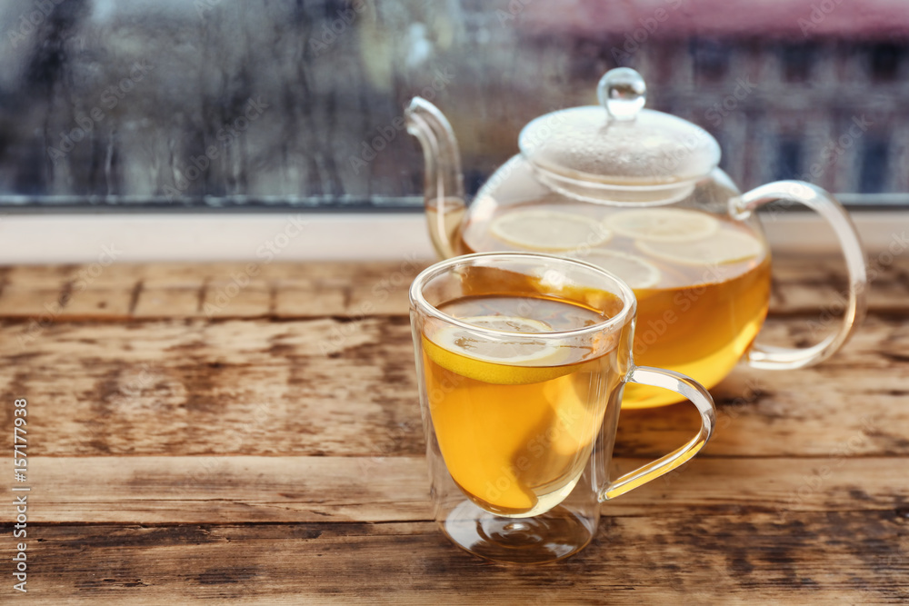 Glass cup of hot tasty drink with teapot on wooden windowsill Glass cup of hot tasty drink with teapot on wooden windowsill