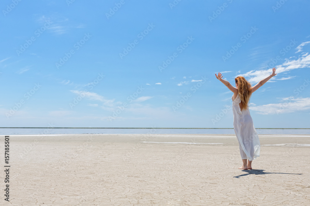 Beautiful young woman in white dress is standing without shoes in the sand on a blue sky background, her hands are raised up. Beautiful young woman in white dress is standing without shoes in the sand on a blue sky background, her hands are raised up.