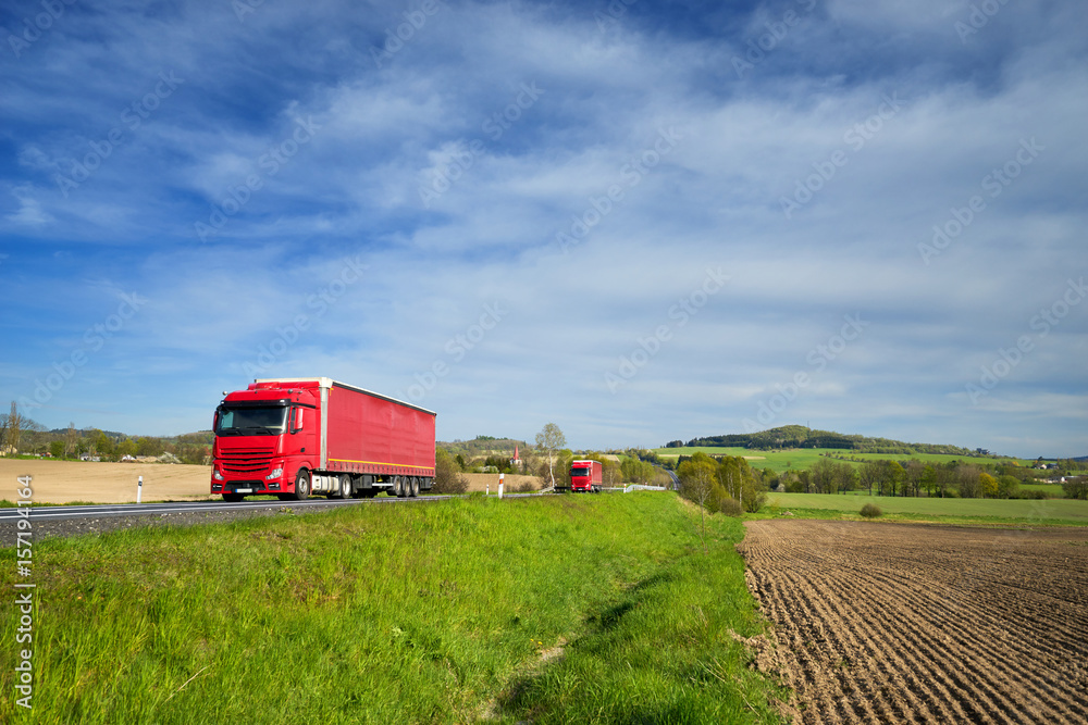 Red trucks driving on the road around a sown field in a rural landscape with a village in the background