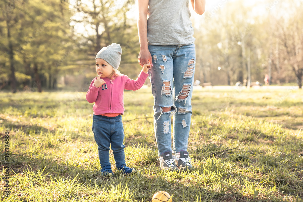 Fototapeta premium Child with mother standing together with holding hands in summer park on grass. Main subject is child. Unrecognizable mother on photo. Concept for togetherness and care.
