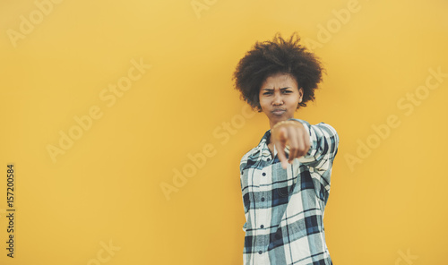 Frowning beautiful afro american teenage girl in checkered shirt standing in front of yellow background and pointing on camera with her finger, with copy space zone for text, logo or your advertising