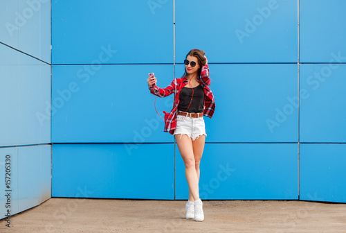 Fashion portrait of pretty smiling hipster girl in sunglasses with smartphone and headphones making selfie against the colorful blue wall. Outdoor