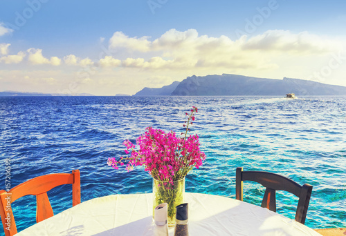 Santorini island, Oia village, Greece. Vase of pink flowers on table with white tablecloth against turquoise water of Aegean sea and mountains in background. Romantic background, nobody scene. © Feel good studio