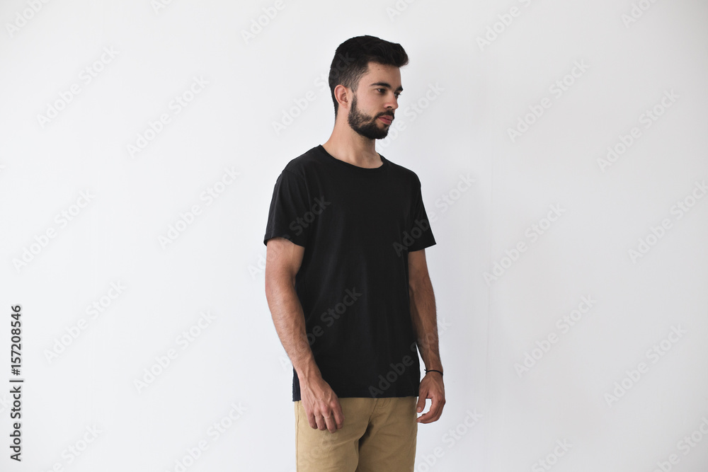 Bearded hipster youngster in studio setting poses in blank black t-shirt and chinos trousers, friendship bracelet softly smiles into the camera, calm and tranquil, on white mockup Bearded hipster youngster in studio setting poses in blank black t-shirt and chinos trousers, friendship bracelet softly smiles into the camera, calm and tranquil, on white mockup