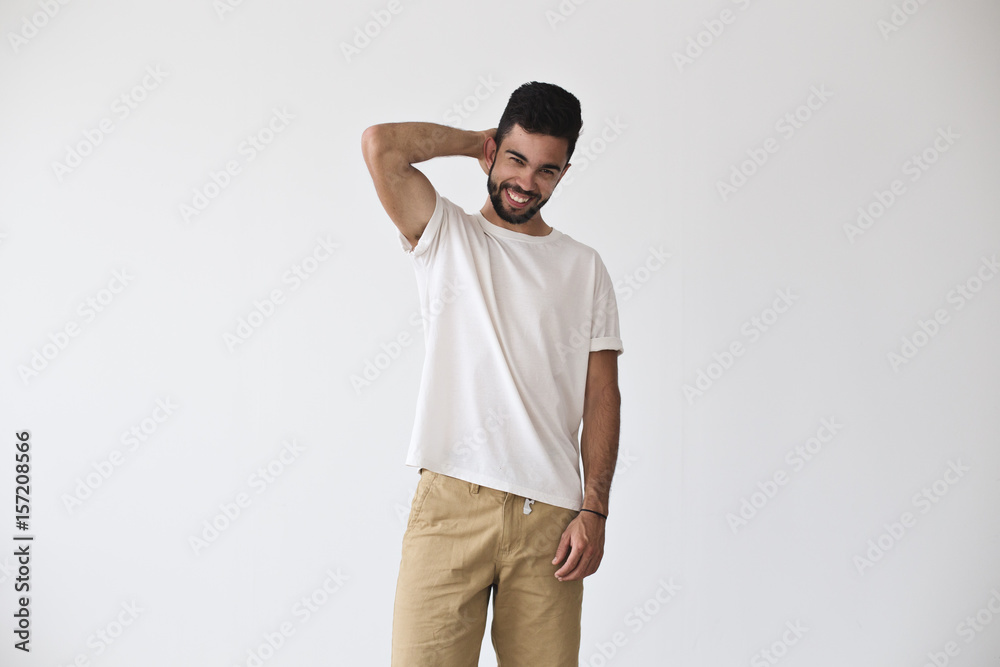 Frontal shot of attractive young hispanic man who chuckles and smiles into camera, in fashion pose in white blank simple tshirt from premium materials, cotton and vyscose Frontal shot of attractive young hispanic man who chuckles and smiles into camera, in fashion pose in white blank simple tshirt from premium materials, cotton and vyscose