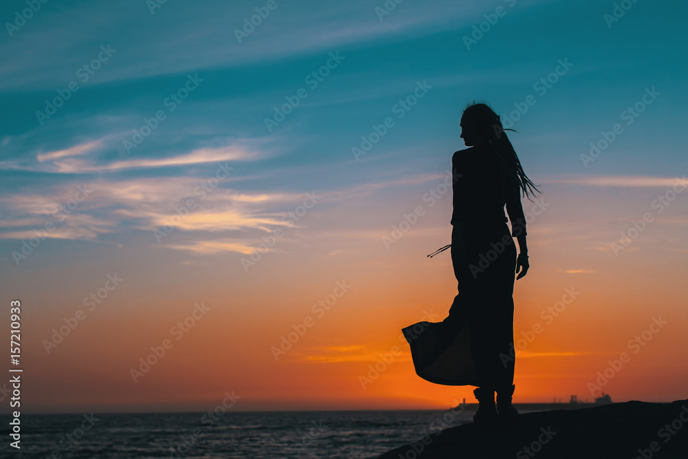 Silhouette of young woman in summer dress standing on a rock and looking to a sea.
