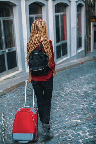 Young woman with blond dreadlocks is on the pavement with a red suitcase. Rear view. Travel and tourism.
