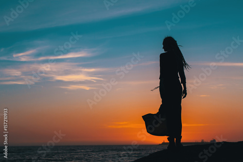 Silhouette of young woman in summer dress standing on a rock and looking to a sea.