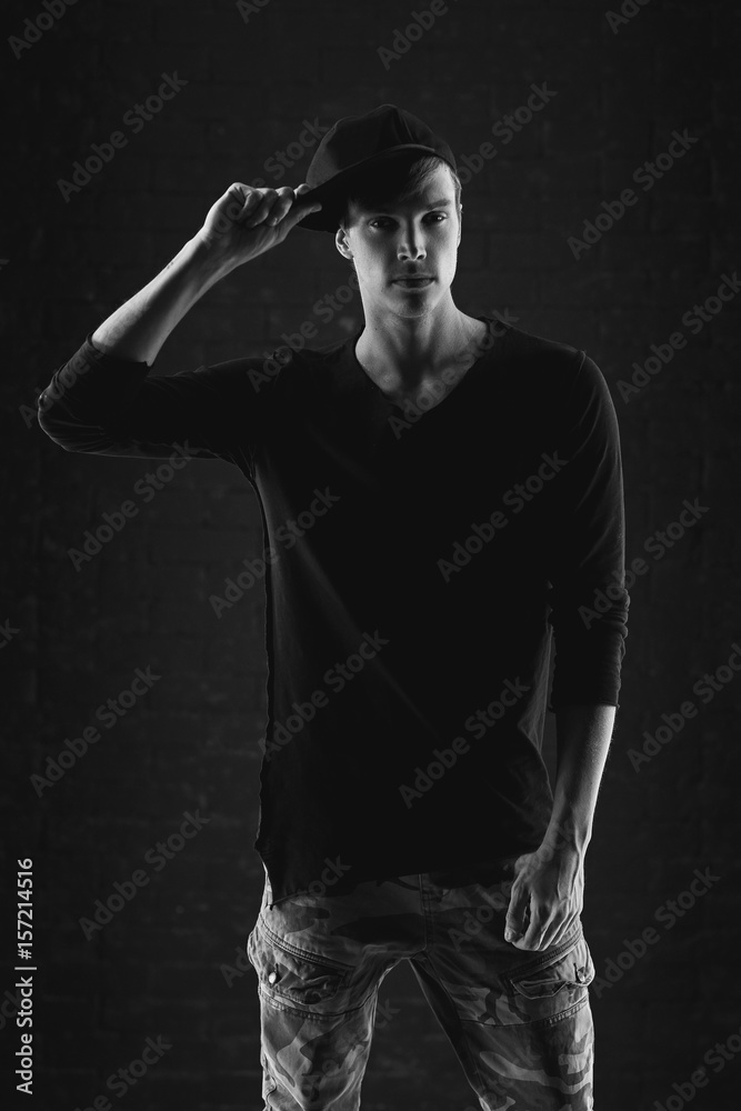 Fashion vogue style portrait of young handsome man in black pullover and cap posing against brick wall background.