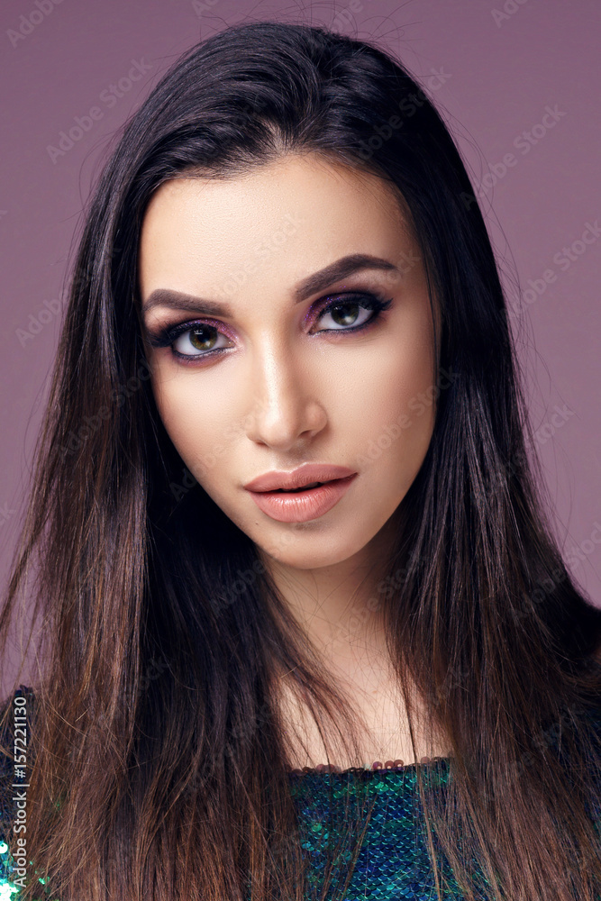 Fototapeta premium Closeup beauty studio fashion portrait of young pretty girl with long brunette dark hair and nakeup posing in shiny green dress with sequins posing against purple background and looking in camera.