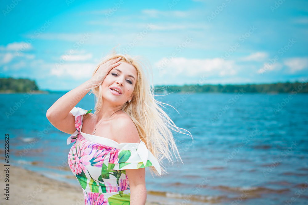 Beautiful blonde hair woman in romantic flowers dress near blue water and sand. Cute happy girl have fun on a nature Beautiful blonde hair woman in romantic flowers dress near blue water and sand. Cute happy girl have fun on a nature